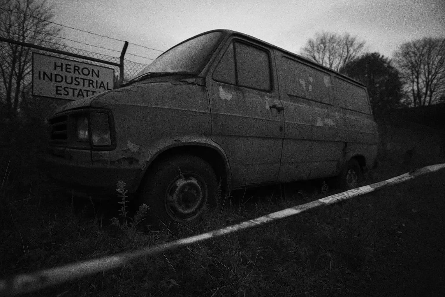 En grå varevogn med afskallet maling står forladt på Heron Industrial Estate, omgivet af tilgroede ukrudt. Politibånd flagrer i nærheden, hvilket antyder dens forbindelse til de uopklarede Hammersmith Nude Murders.